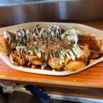 Delicious takoyaki balls drizzled with sauce and topped with bonito flakes, served in a wooden tray at a street food stall.