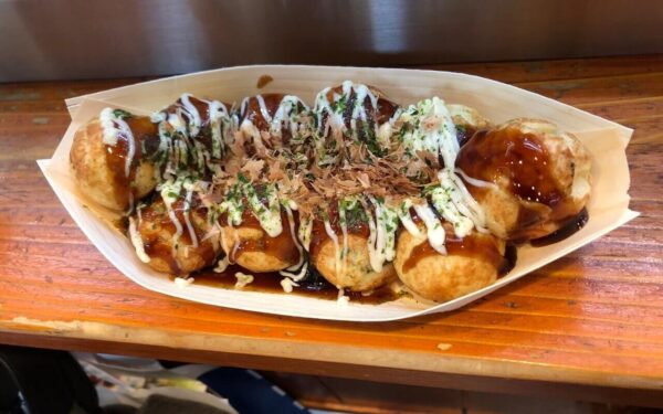 Delicious takoyaki balls drizzled with sauce and topped with bonito flakes, served in a wooden tray at a street food stall.