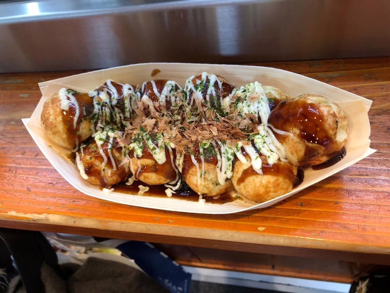 Delicious takoyaki balls drizzled with sauce and topped with bonito flakes, served in a wooden tray at a street food stall.