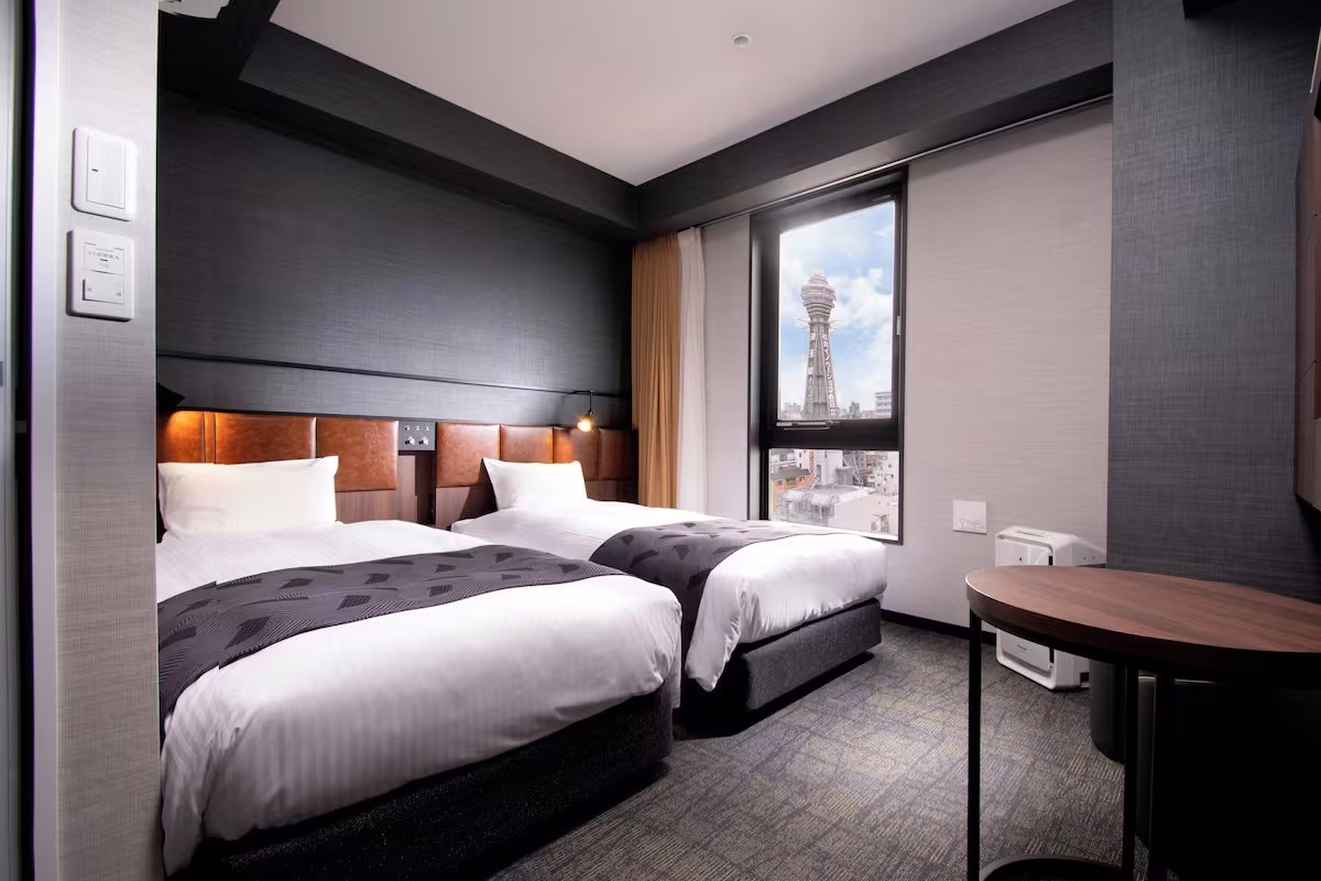 Modern hotel room with twin beds, offering a view of a landmark tower through a large window, perfect for travelers.