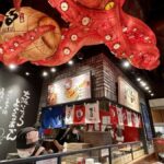 Vibrant restaurant interior with an octopus lantern and chefs preparing traditional Japanese cuisine in a lively atmosphere.