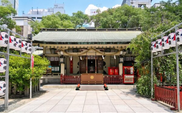 Serene shrine entrance with traditional lanterns and red offerings, showcasing Japanese culture amid urban surroundings.