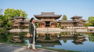 Kannaya Nareswari in a green coat walks by a serene pond reflecting a traditional Japanese temple.