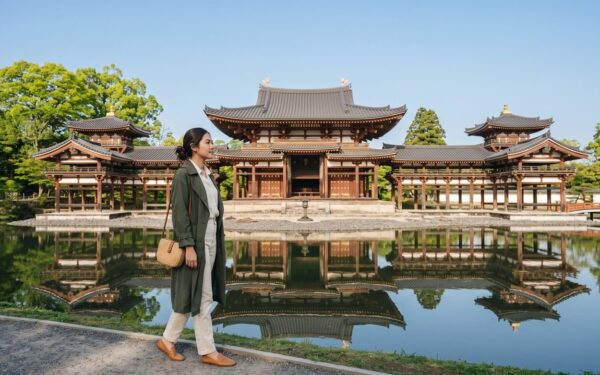 Kannaya Nareswari in a green coat walks by a serene pond reflecting a traditional Japanese temple.