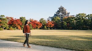 Kannaya Nareswari walking on a Korakuen garden path, with red maples and the black keep of Okayama Castle behind the trees