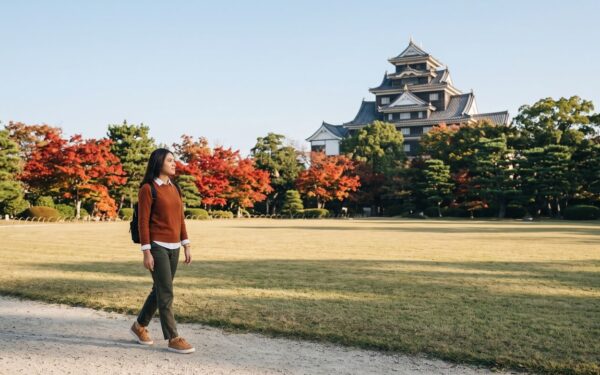 Kannaya Nareswari walking on a Korakuen garden path, with red maples and the black keep of Okayama Castle behind the trees
