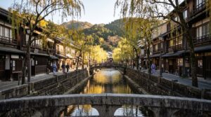 A picturesque canal runs through a traditional Japanese town with wooden buildings and weeping willow trees.