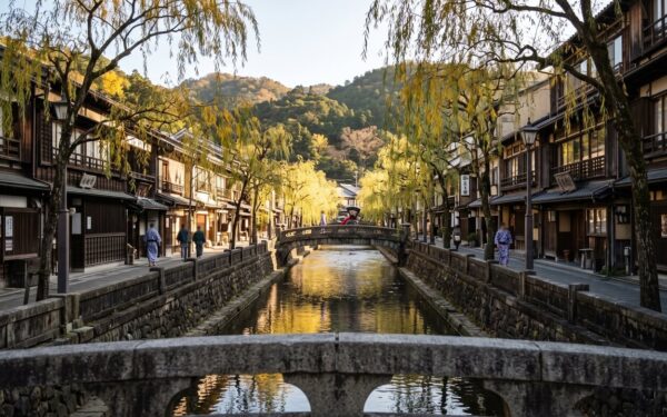 A picturesque canal runs through a traditional Japanese town with wooden buildings and weeping willow trees.