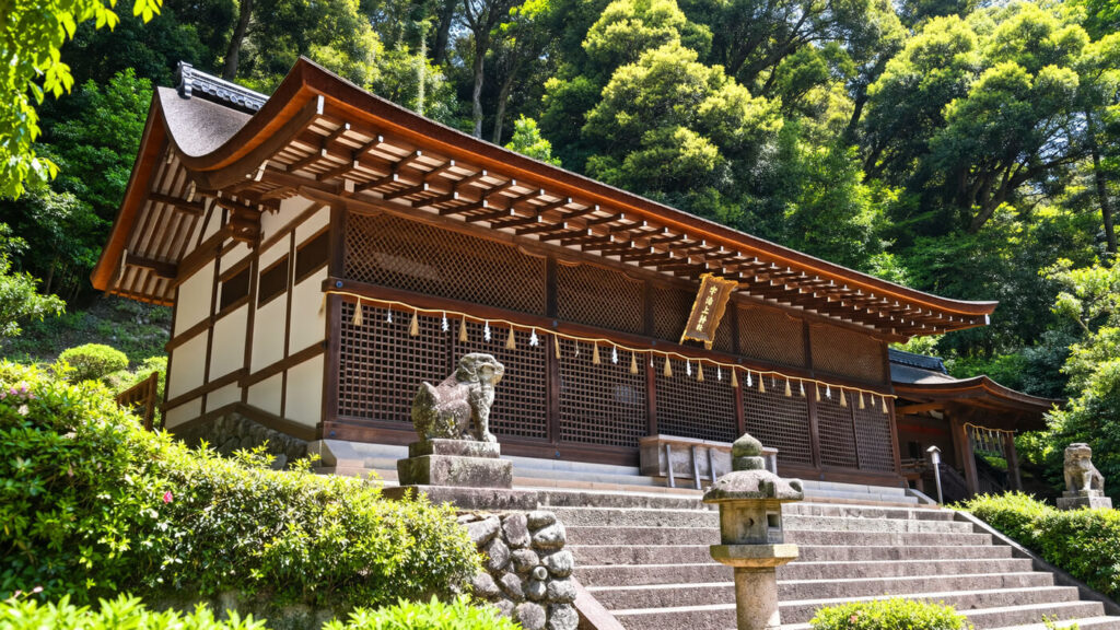 A stone lion statue guards the stone steps leading to a traditional Japanese shrine nestled among lush green trees.